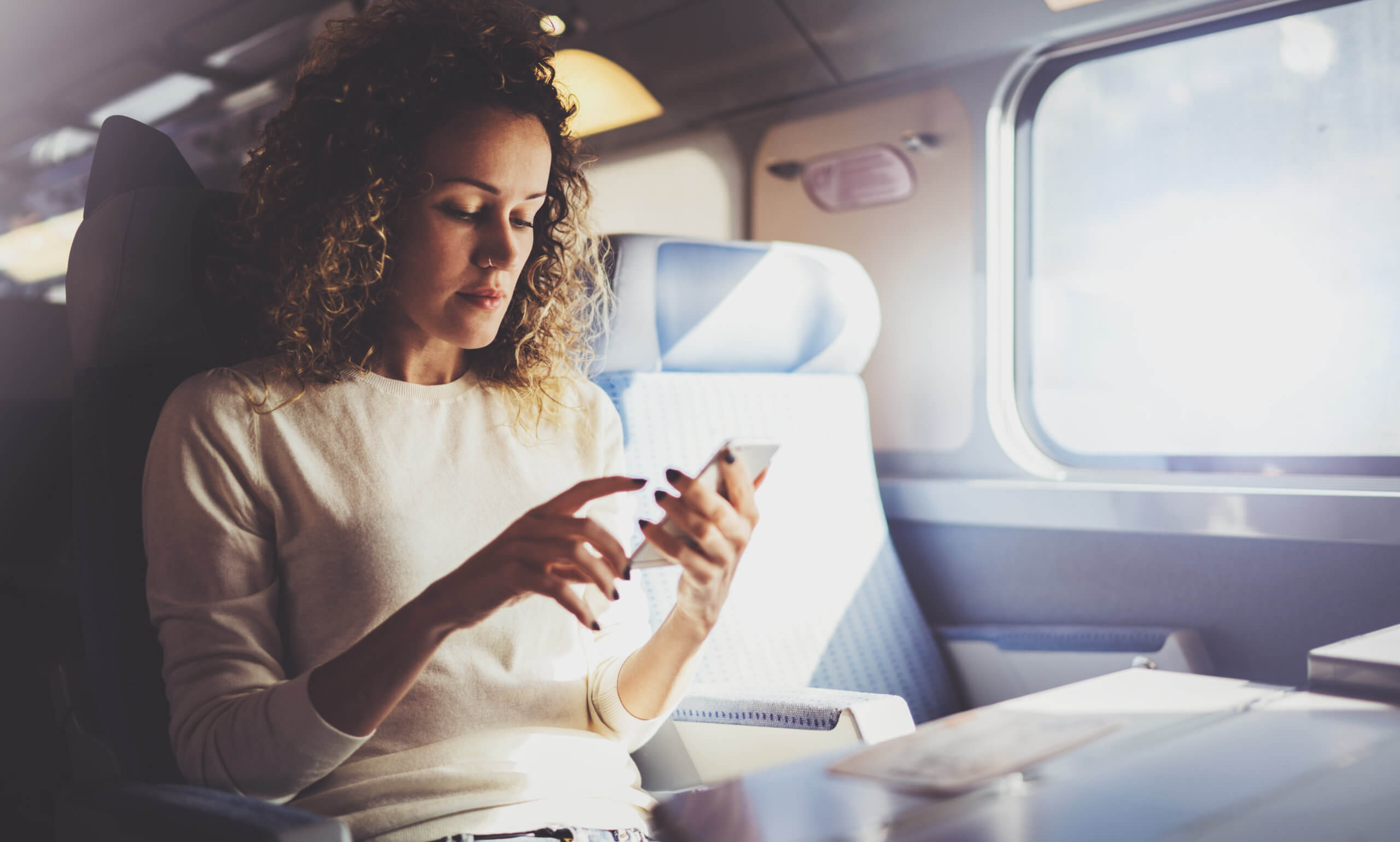 Curly haired, mid thirties ethnically ambigious woman looking at her smart phone while sitting on a train; sun is streaming through the window. stream ctv